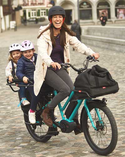 Frau mit 2 Kindern auf Gepäckträger fährt mit Lasten E Bike durch Zürich